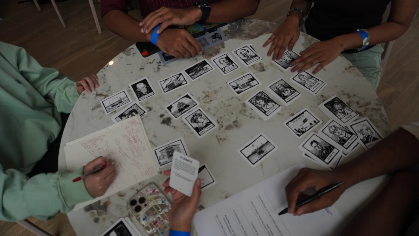 Overhead view of a white table covered in black-and-white photo cards, with four sets of hands holding cards and pens
