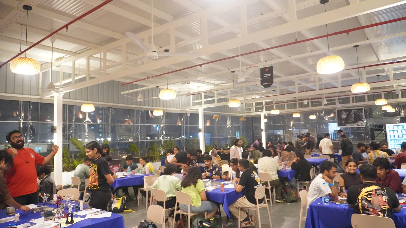 The hall at night with warm pendant lights on and every table full of players seated over their games