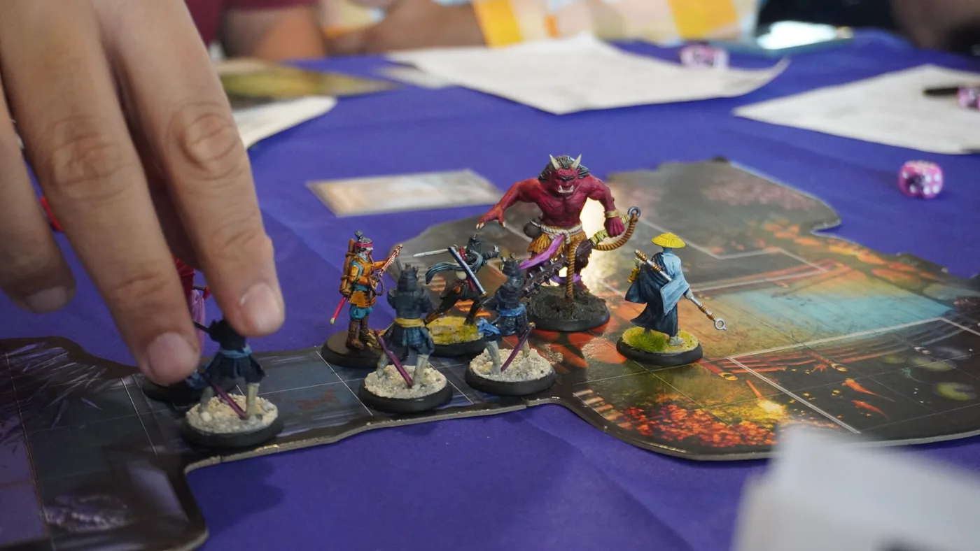 Hand-painted samurai miniatures facing a red demon figure on a printed battle map, a player's hand moving one of them